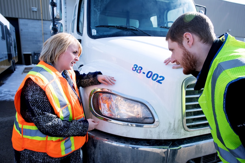 Two people, wearing safety vests, examine the headlight of a white truck with the number 88-002 on it. The scene appears to be outdoors in an industrial area. The woman is pointing at the headlight while the man looks closely at it.