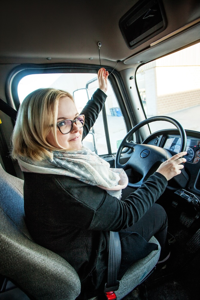 A person with blonde hair and glasses is sitting in the driver's seat of a truck, holding the steering wheel with one hand and pulling a cord above with the other hand. The truck's interior, including the dashboard, is visible.