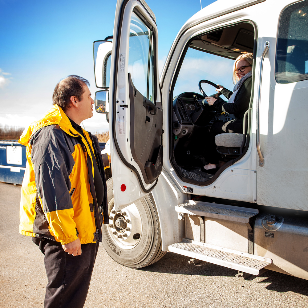 A man in a yellow and black jacket stands outside the open door of a white truck, speaking to a woman sitting in the driver's seat. They appear to be conversing, with both looking towards each other. The scene is set outdoors on a clear day.