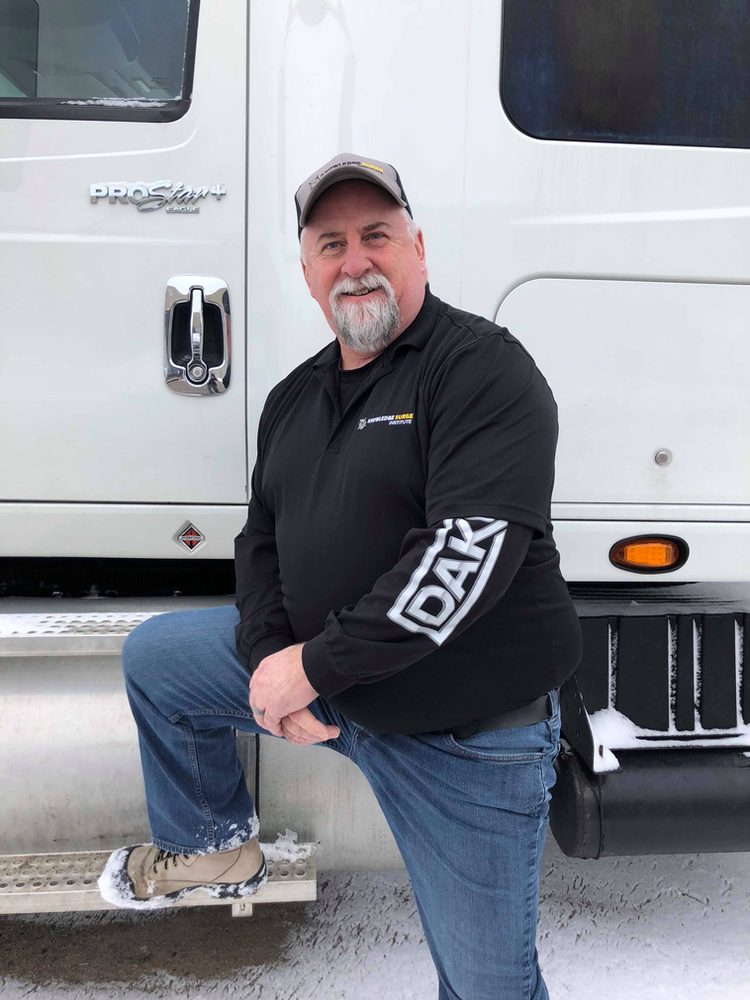 A man with a white beard and mustache, wearing a black cap, black long-sleeve shirt, jeans, and beige boots, stands with one foot on the step of a large white truck. He is looking directly at the camera, with a snowy ground visible around him.