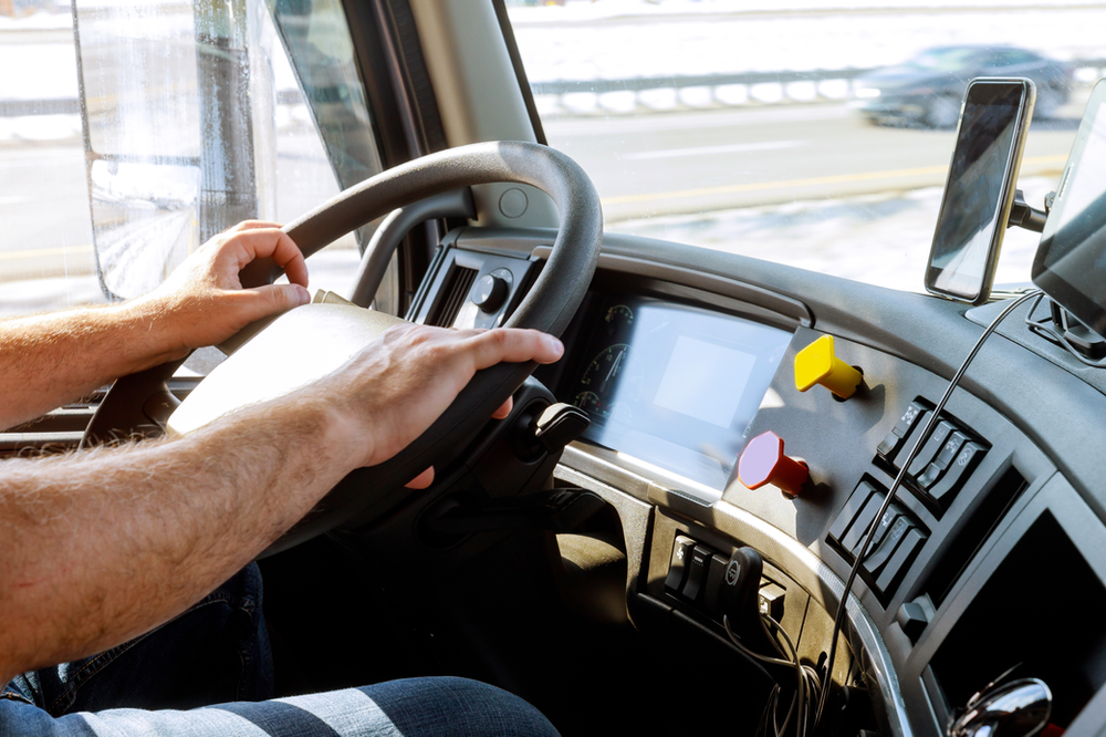 A person is driving a vehicle, focusing on the road ahead. The interior of the vehicle shows a steering wheel, a dashboard, and a mounted smartphone on the right side. The driver's hands are on the wheel, and an arm rest is visible.