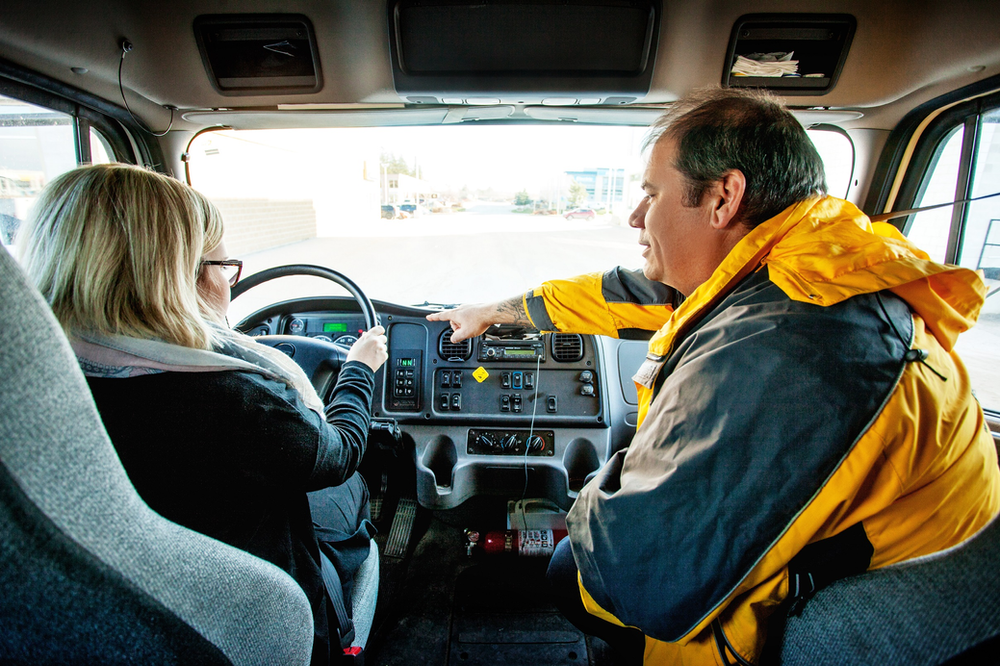 A man in a yellow jacket points at the dashboard of a truck while talking to a woman sitting in the driver's seat. The woman is looking at where he is pointing. The interior of the truck cab is visible, with the steering wheel and various controls in view.