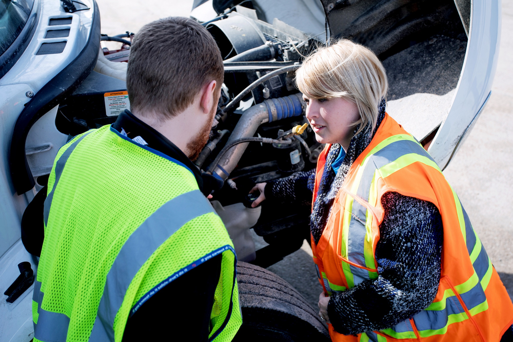 Two people wearing high-visibility vests inspect the engine of a large truck. The individual on the left, wearing a yellow vest, appears to be explaining something, while the person on the right, in an orange vest, listens attentively. The truck hood is open.