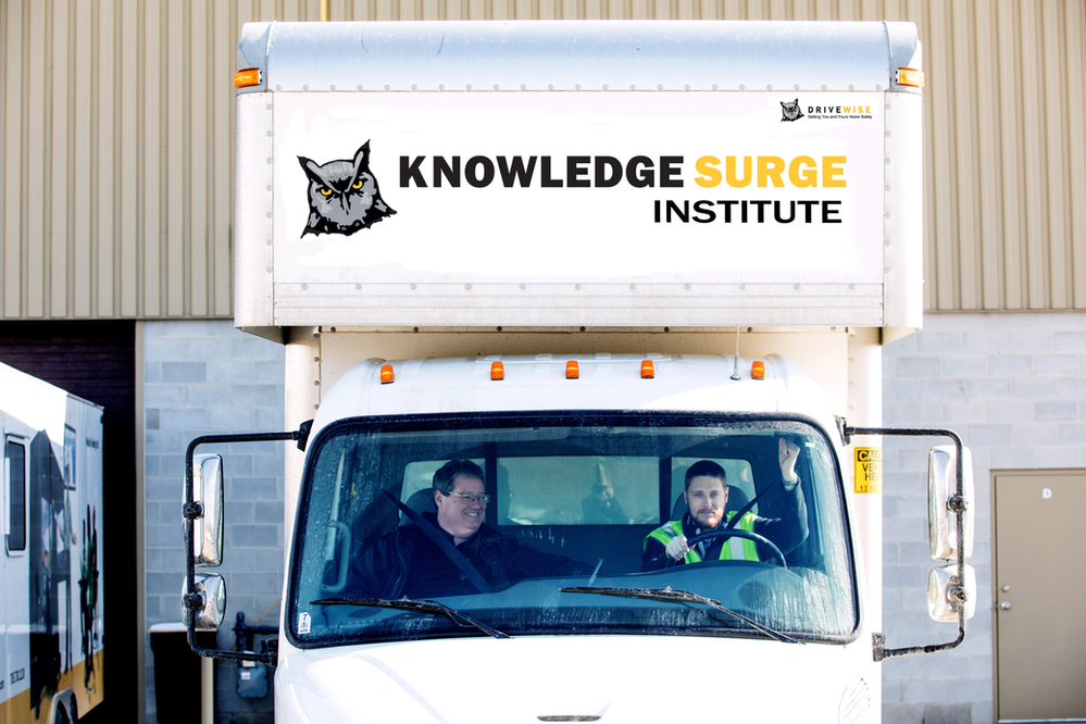 A truck with the "Knowledge Surge Institute" logo on its front box is parked. Two people are seated inside the truck, smiling. The truck is in front of a building with a beige exterior and a brown garage door. Another truck is visible in the background on the left.