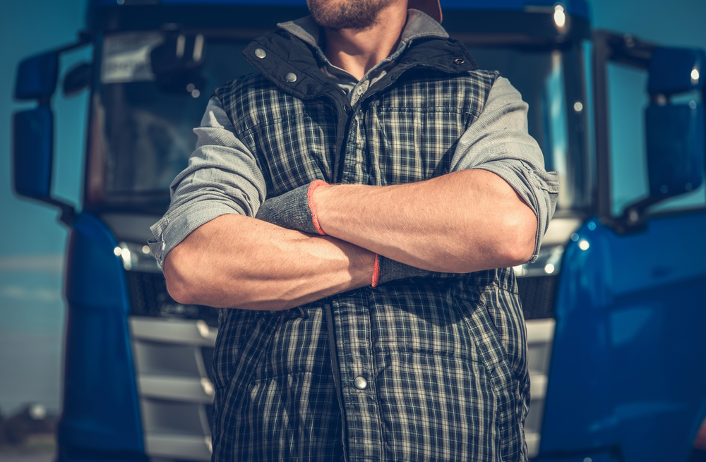 A man crossing his arms in front of a blue truck. He is wearing a plaid vest over a long-sleeve shirt and a baseball cap. His face is not visible in the image.