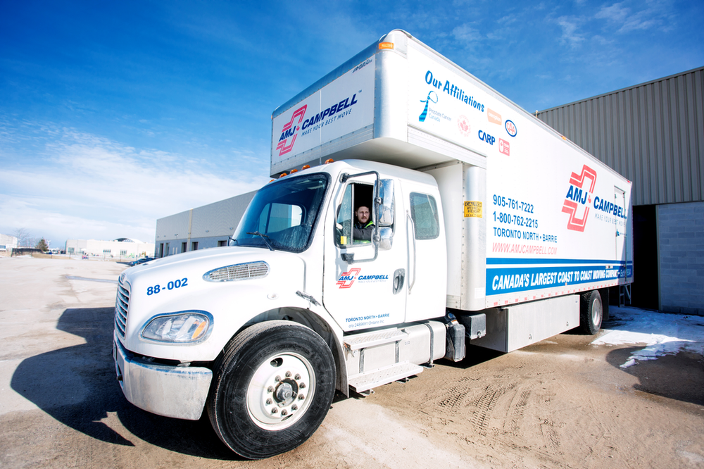 A white delivery truck with the AJ Campbell logo and "Canada's largest coast to coast movers" on the side is parked outdoors. A person is visible through the driver's window, giving a thumbs-up gesture. The background shows a building and a clear sky.